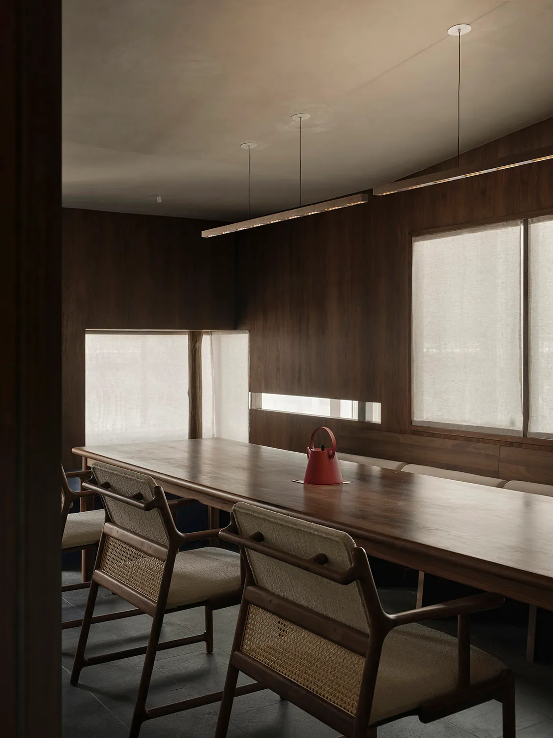 Modern dining room featuring a long wooden table and chairs with a red kettle on the table, large windows with sheer curtains