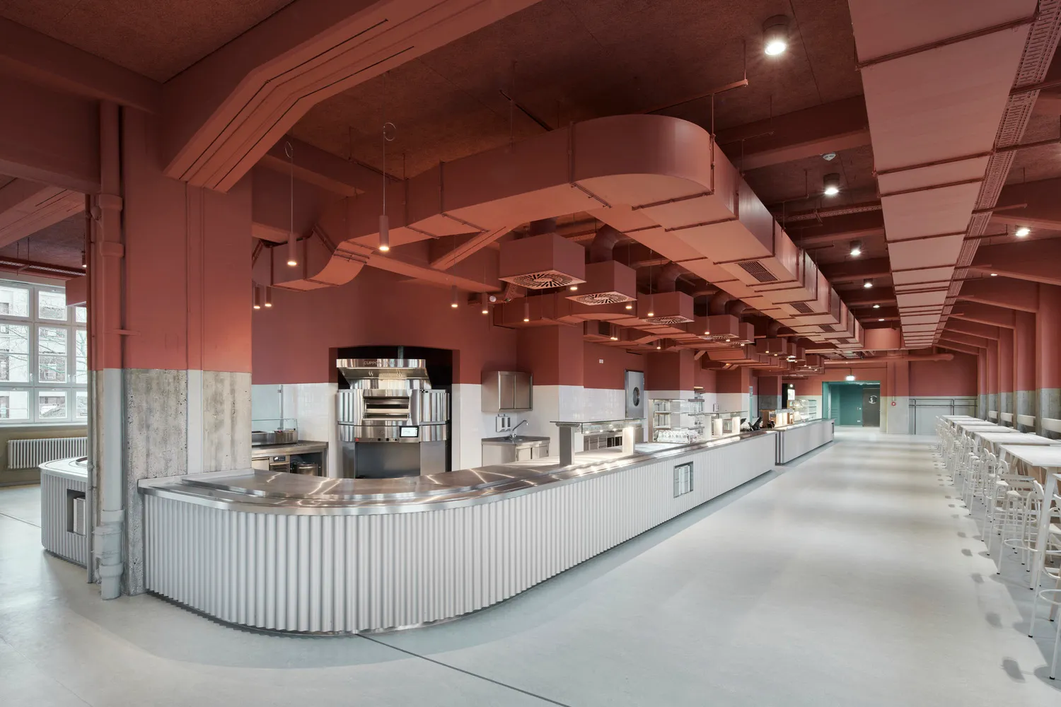 An industrial-style cafeteria with a long metal counter, white tile backsplash, and exposed ventilation systems.