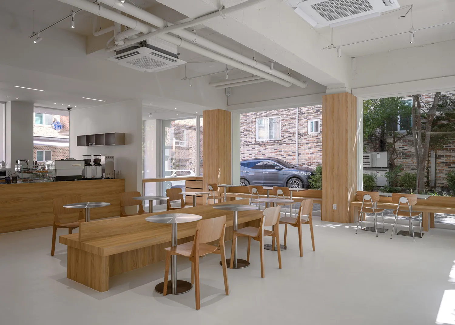 Modern cafe interior with wooden tables and chairs, white walls, and large windows.