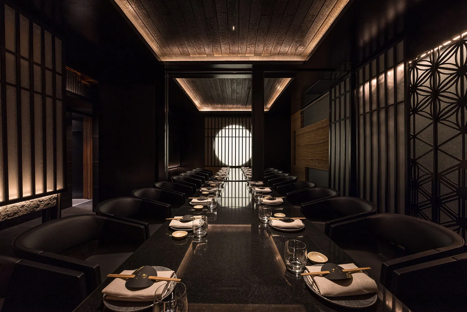 A modern restaurant interior with black furniture, featuring a long dining table set with white plates, chopsticks, and glass