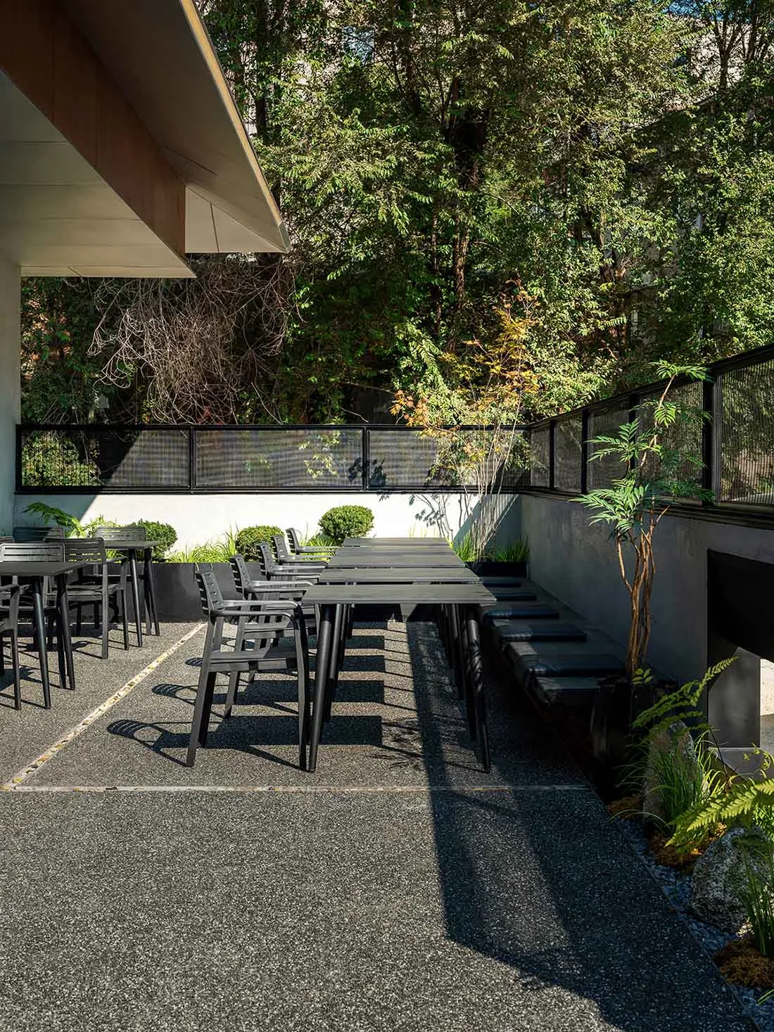 Modern minimalist outdoor dining area with concrete flooring, gray table, and black chairs surrounded by lush greenery and a