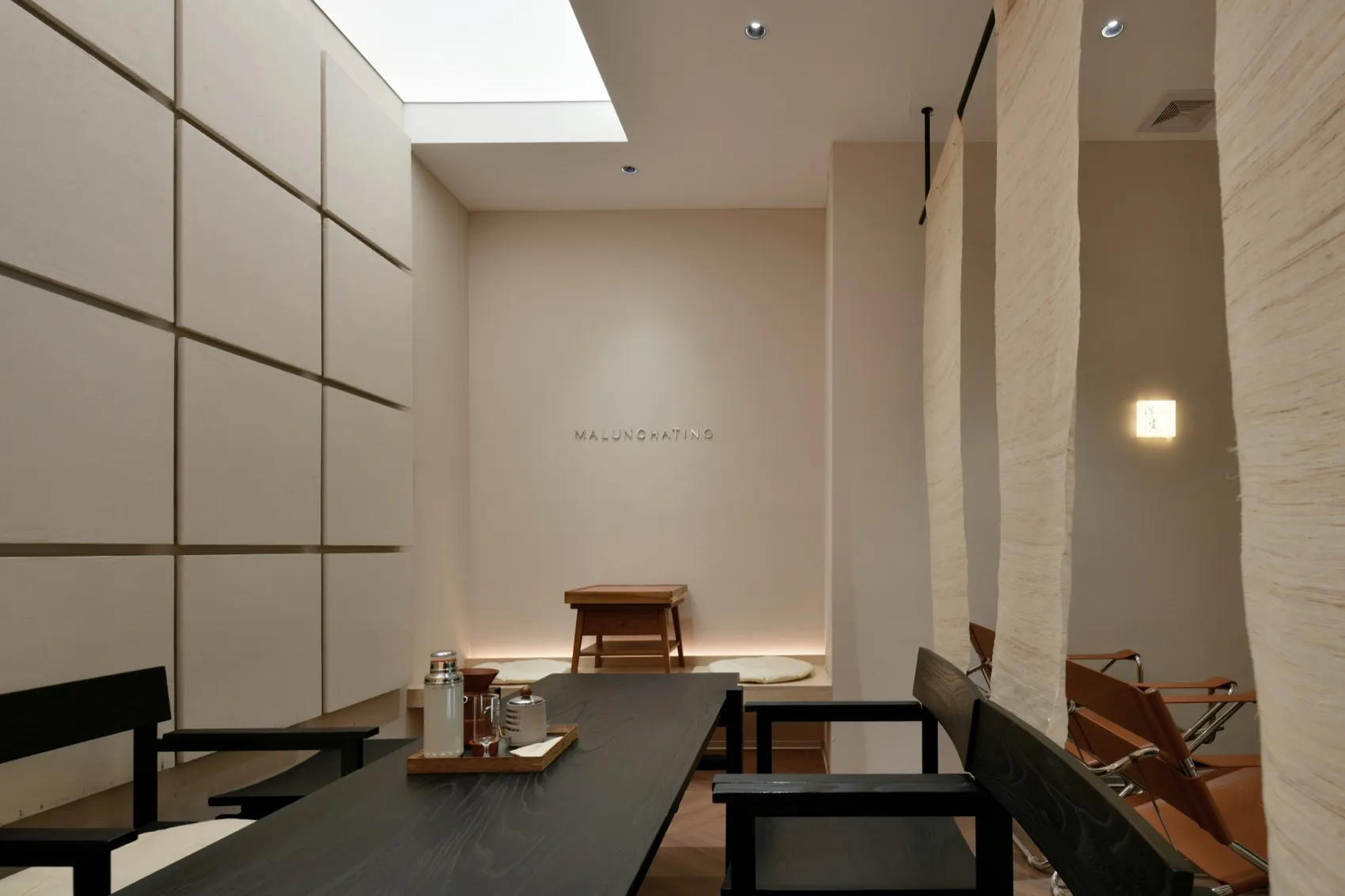 A minimalist restaurant interior featuring a wooden dining table and chairs, soft lighting, and a skylight.