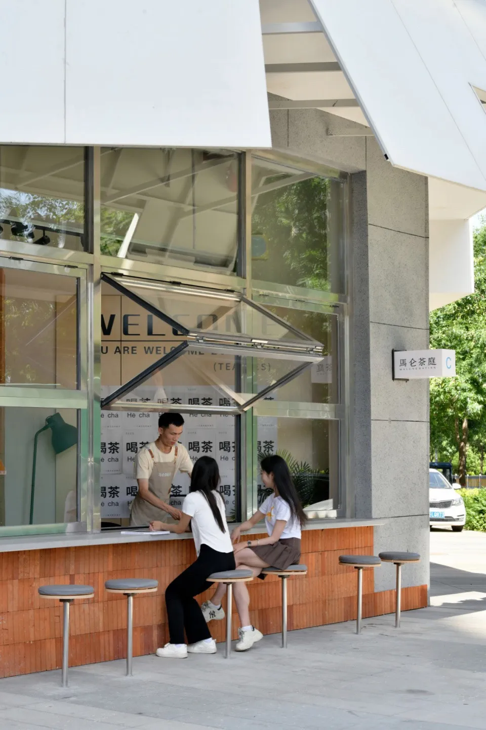 Two young women sitting outside a modern coffee shop with a glass storefront and a wooden countertop.