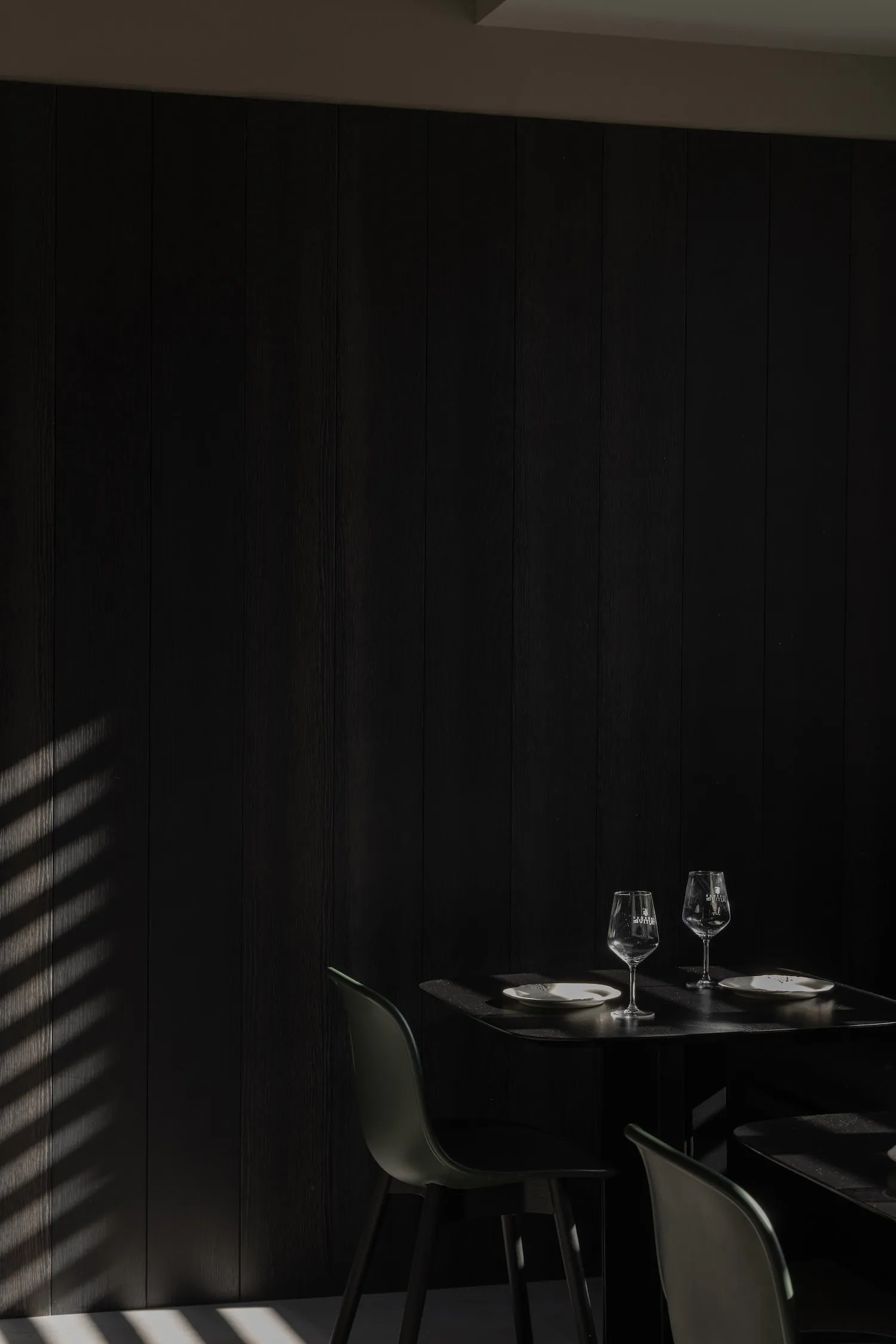 A modern dining room with dark wood walls and a small black table set for two, featuring two wine glasses and plates.