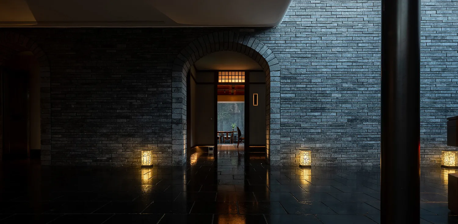 A dimly lit lobby with a dark brick wall and floor, featuring a table and chairs in the distance.