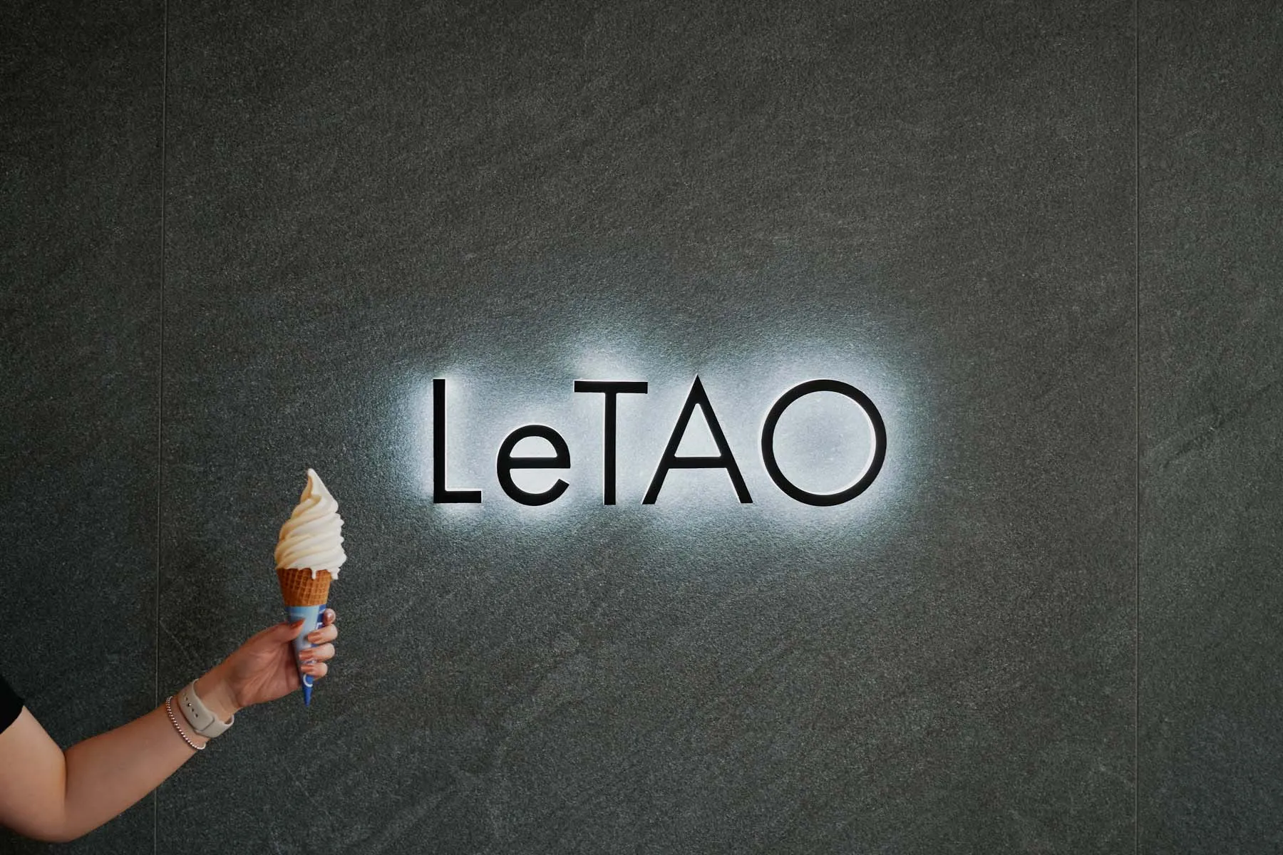一隻手拿著冰淇淋甜筒,站在灰色大理石招牌前,背景有「LeTAO」燈光照亮。