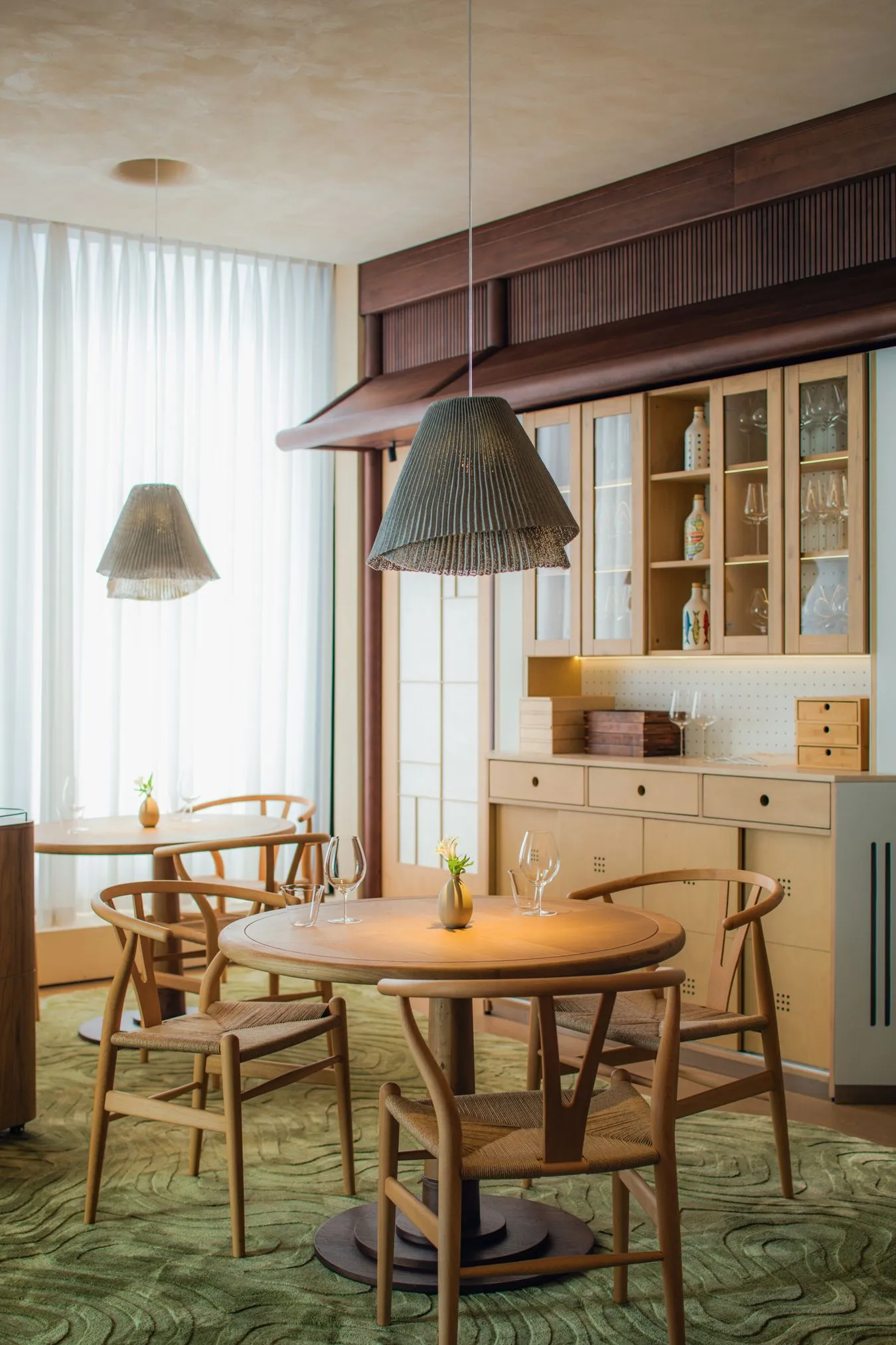A modern restaurant interior with a wooden dining table and chairs, wooden counter, and hanging pendant lights.