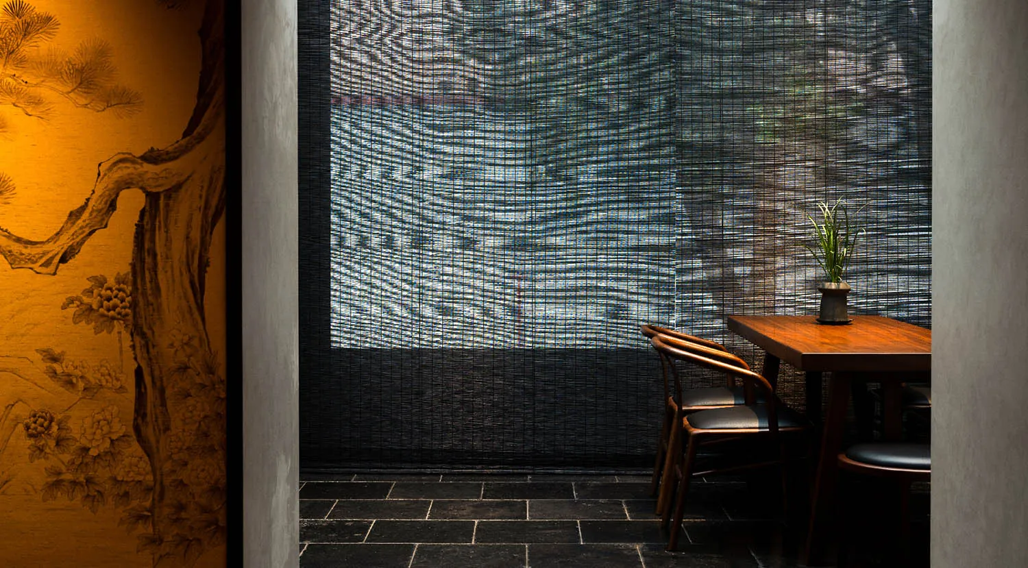 A minimalist restaurant interior featuring a wooden table, black cushions, and a bamboo shade covering a large window.