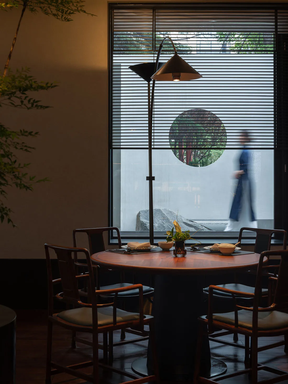 A modern restaurant interior featuring wooden tables, metallic floor lamp, and plants.