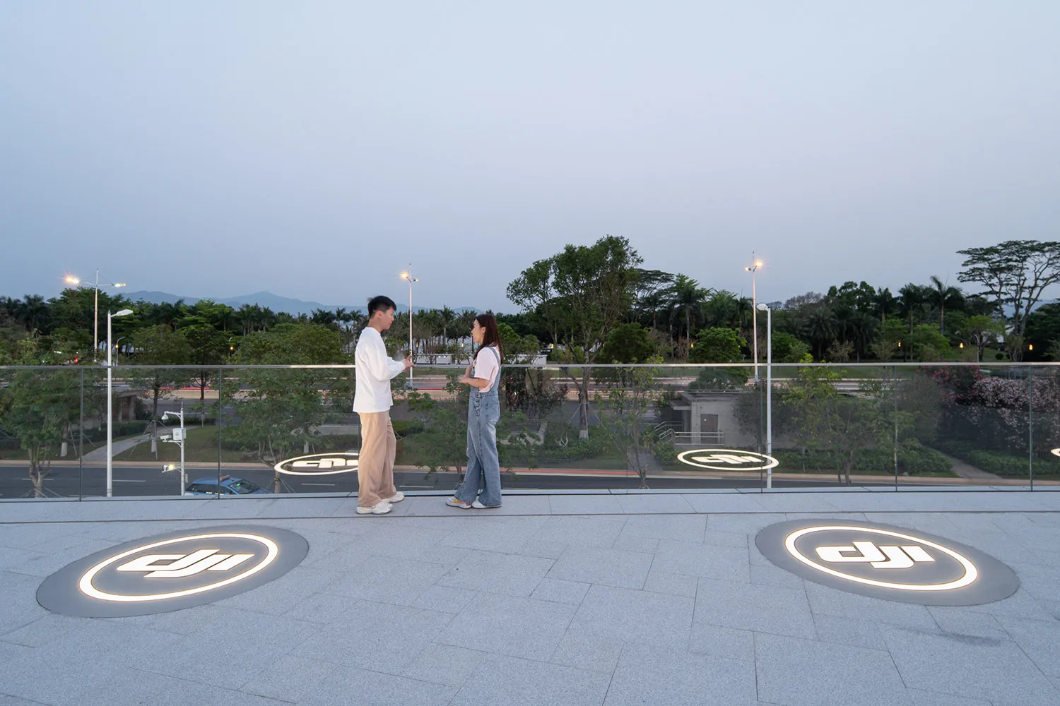 A couple standing on a glass platform overlooking a park at dusk.