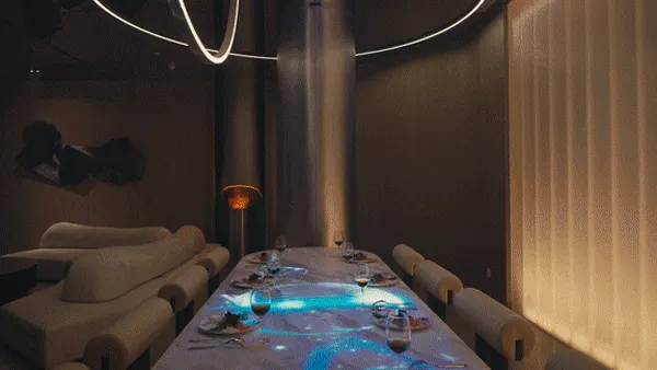 一間空蕩的工業風餐廳,長餐桌已擺設餐具,燈光昏黃。