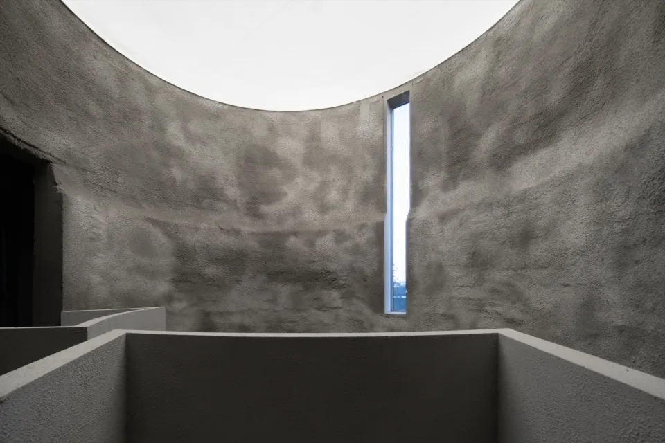 An image depicting a minimalist concrete interior design with a large circular skylight and a tall, narrow window.