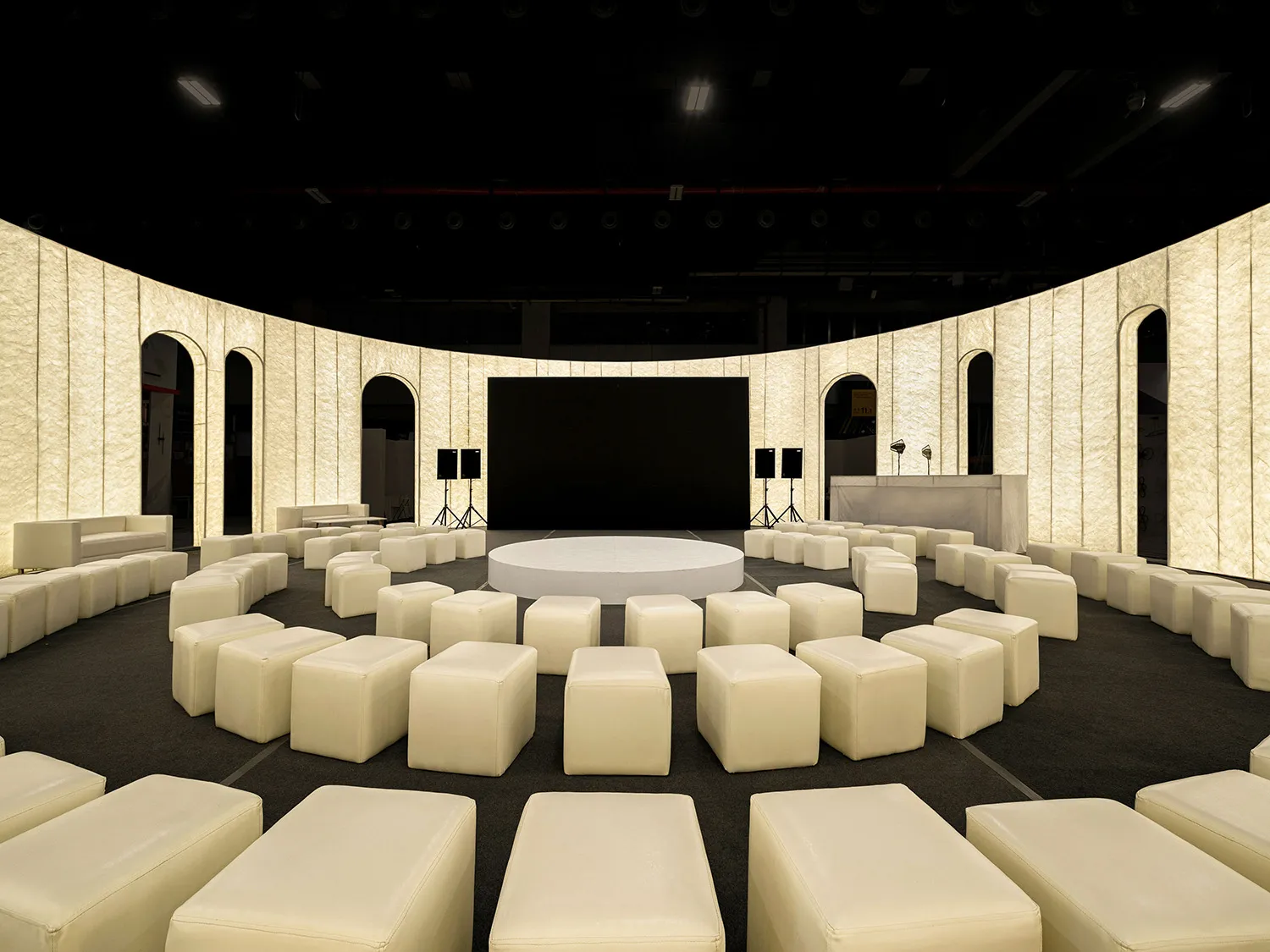 Circular arrangement of fabric-covered seating in a conference center with a stage and media equipment.