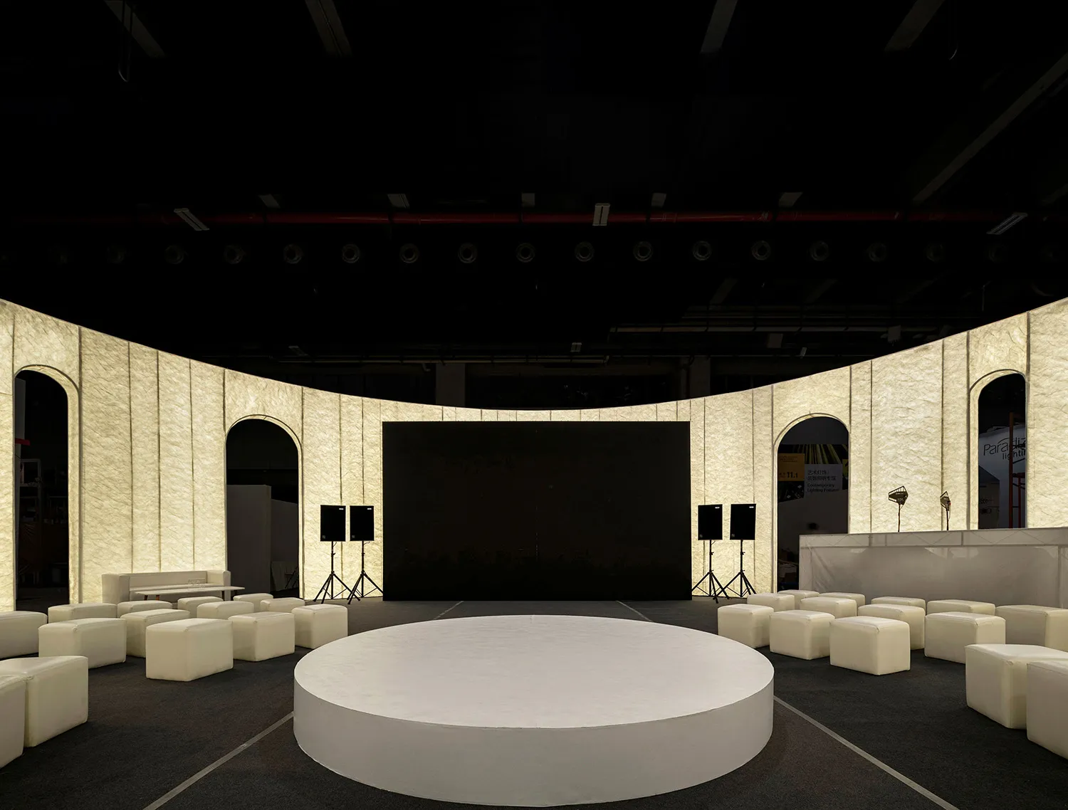 A minimalist conference room with a round, white platform at the center, surrounded by upholstered seating and concrete walls
