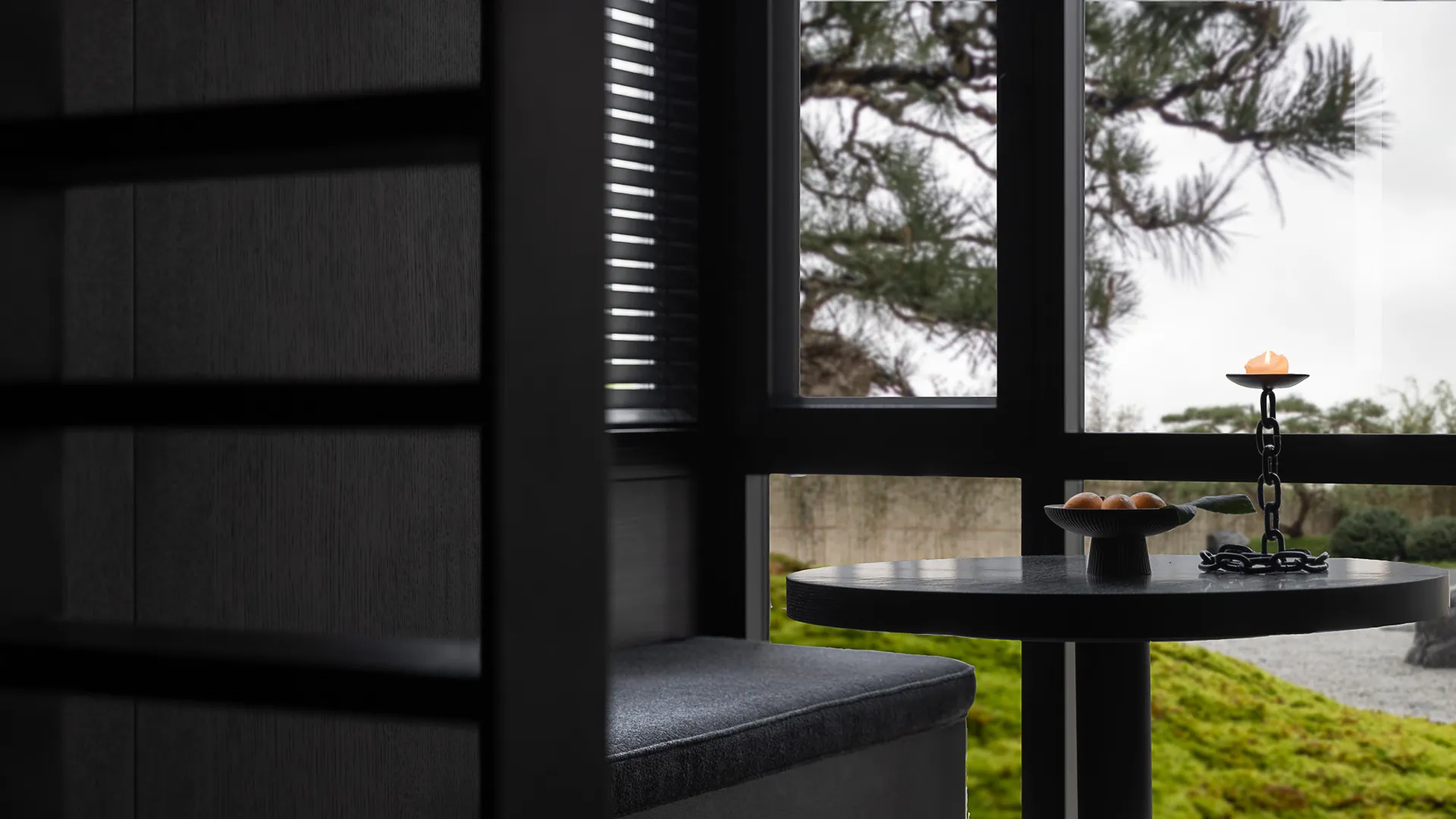A modern interior space featuring a small table with a candle holder and fruit bowl, overlooking a serene garden with trees.