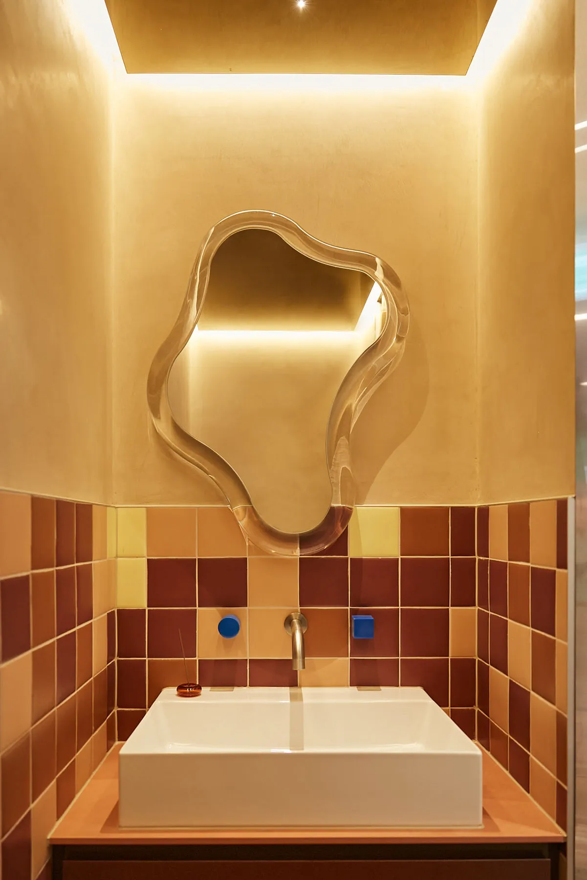 A minimalist bathroom featuring ceramic tiles with a mix of earthy tones and a unique, abstract-shaped mirror.