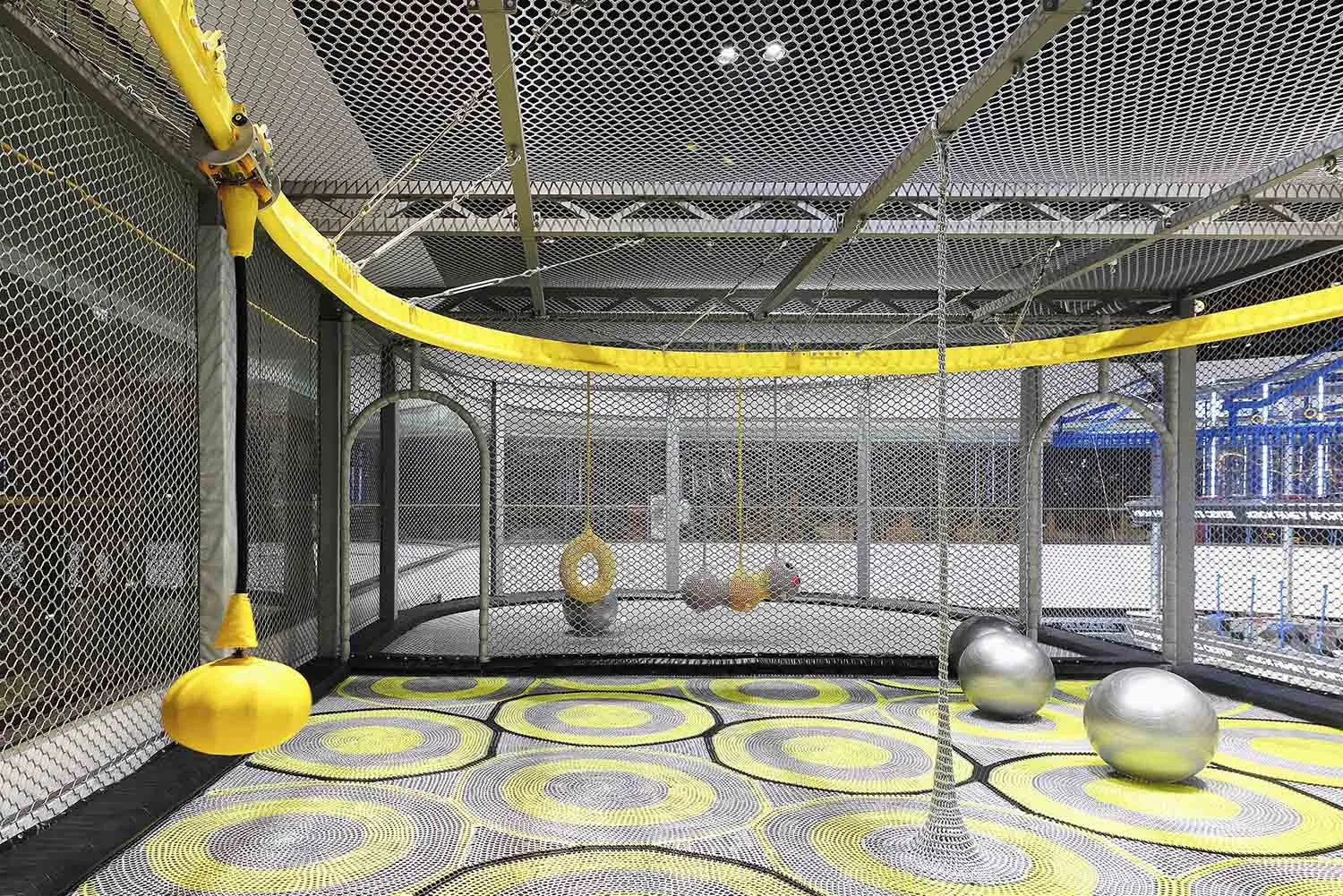 Playground interior with netted walls, metal poles, and suspended balls inside a large, enclosed space with patterned floorin