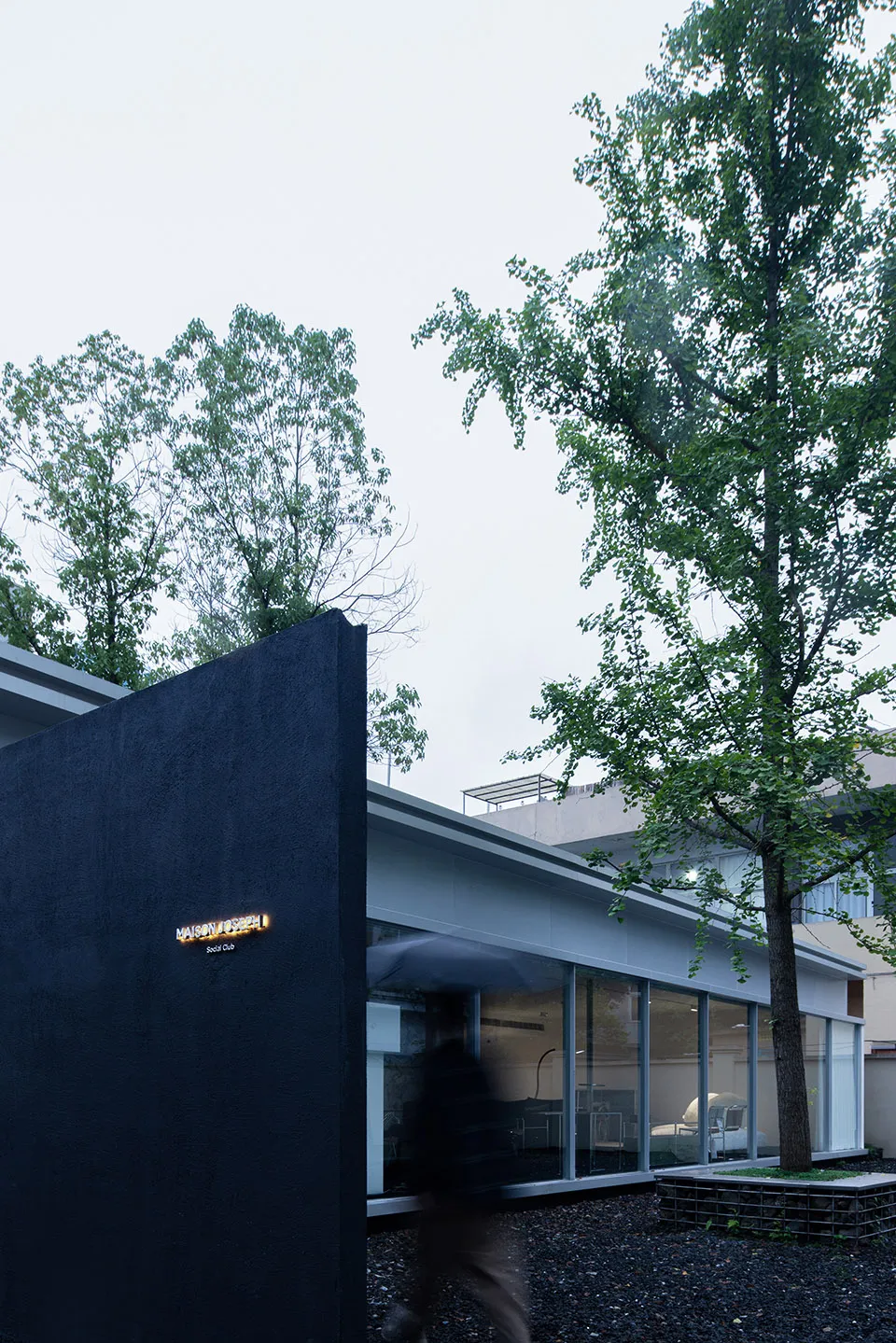 A modern social club with a black wall, glass windows, and steel accents, surrounded by green trees and gravel.