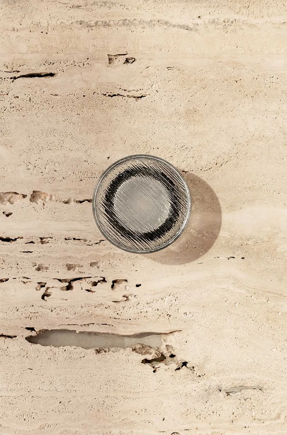 A minimalist glass coaster with a dark circular design on a textured, beige stone surface.