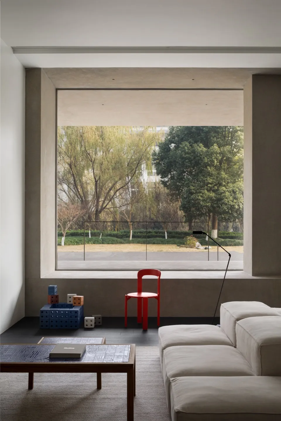 A modern living room featuring concrete walls and floor, with a red chair, white sofa, and a large window overlooking a green