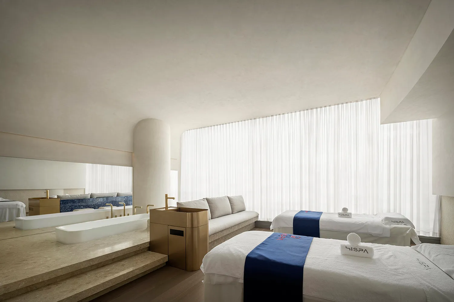 A serene spa room with white cloth textiles on stone flooring, glass walls, and a large window.