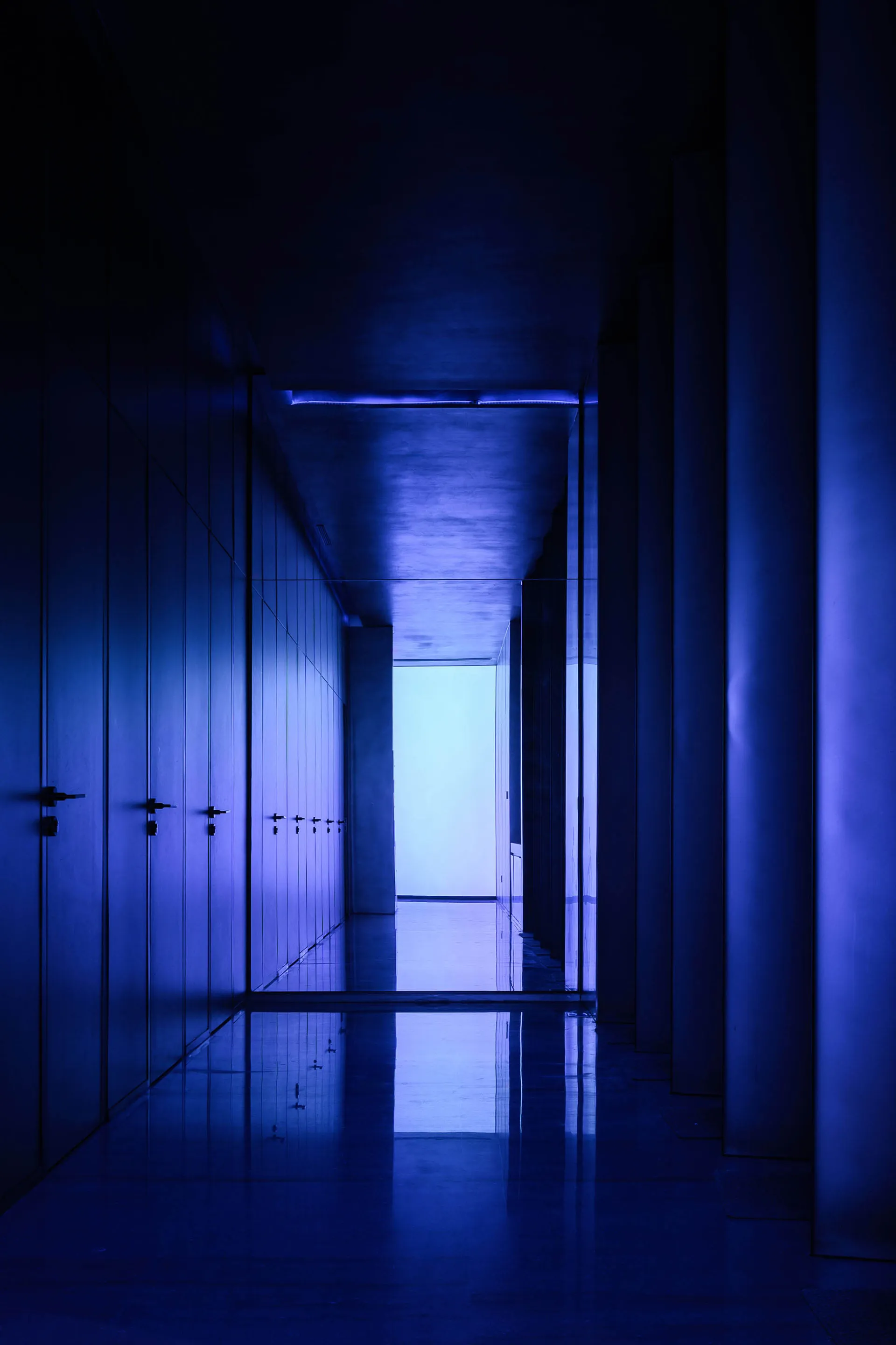 A long, narrow corridor with walls illuminated in a single, deep blue hue, extending towards a bright endpoint.