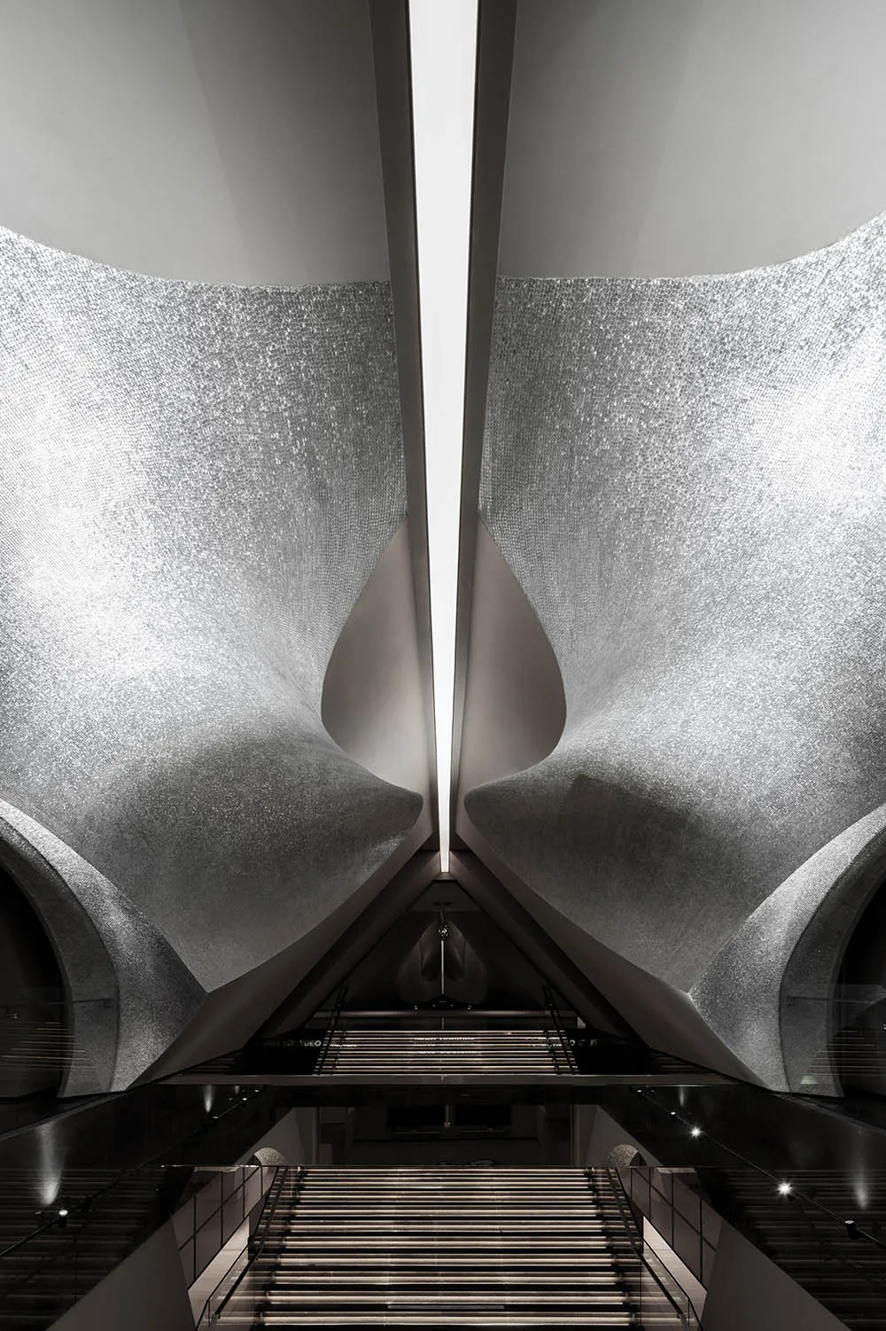 A futuristic, spacious transportation station with metallic walls reflecting light and a staircase leading downwards.