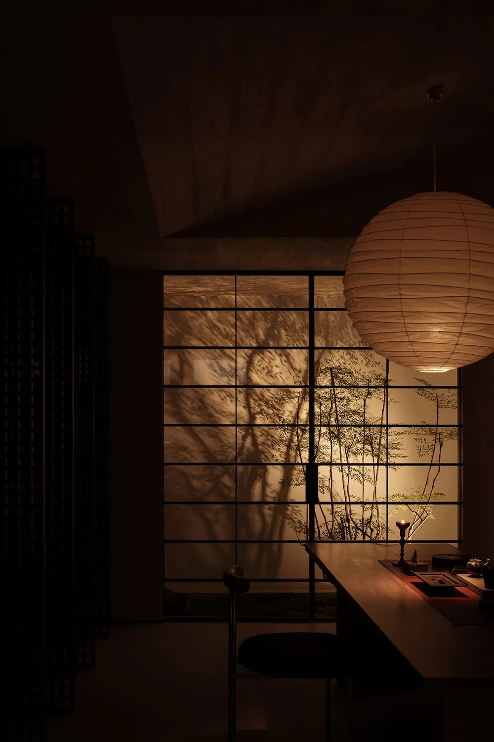 A serene living room with a paper lampshade hanging above, casting shadows on the concrete wall, illuminated by the warm glow