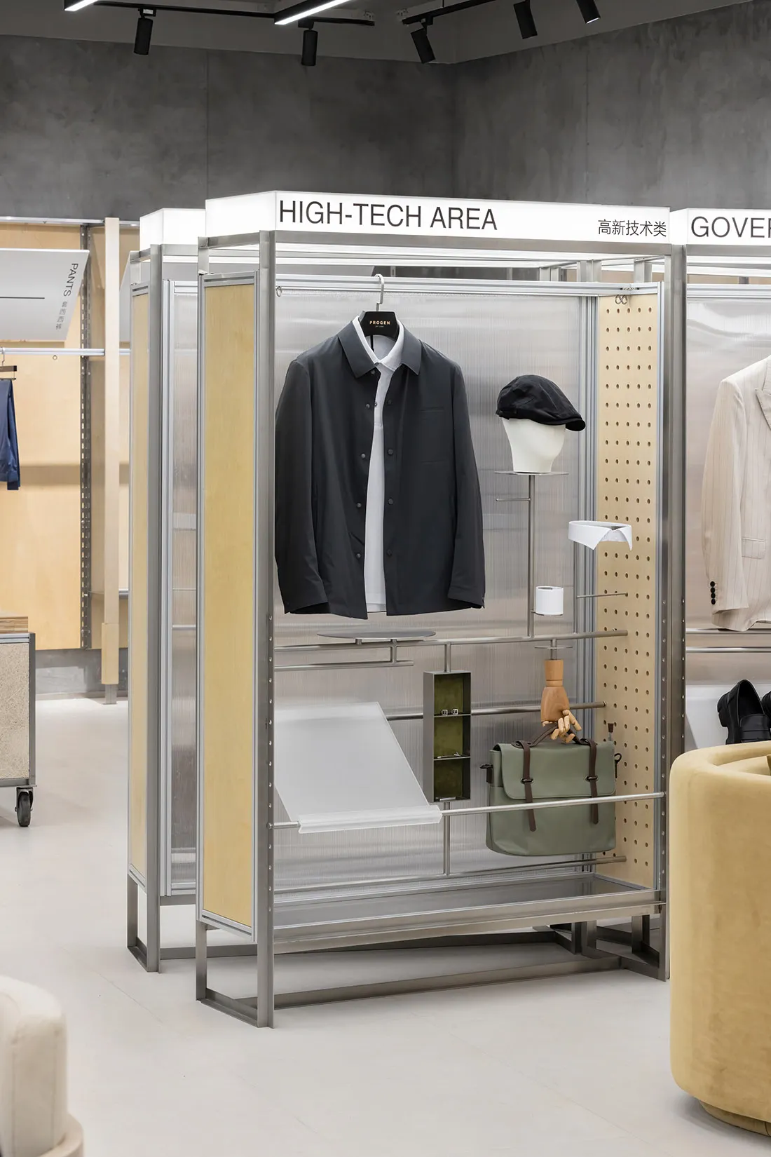 A high-tech clothing shop interior displaying a black jacket, cap, and green bag on a metallic rack with wooden backing.
