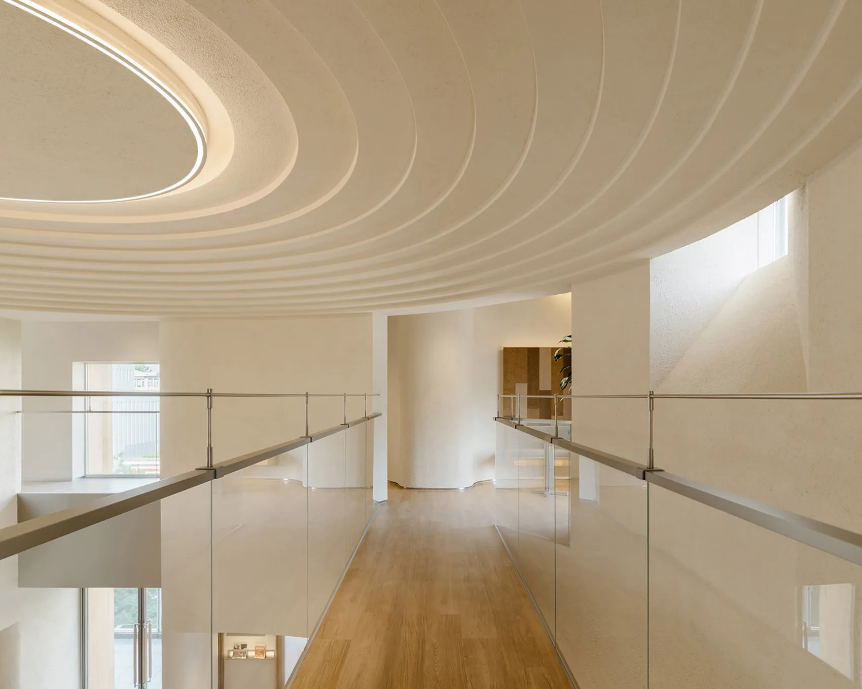 A modern corridor with a glass ceiling and wooden floor, featuring sleek railings and textured walls.