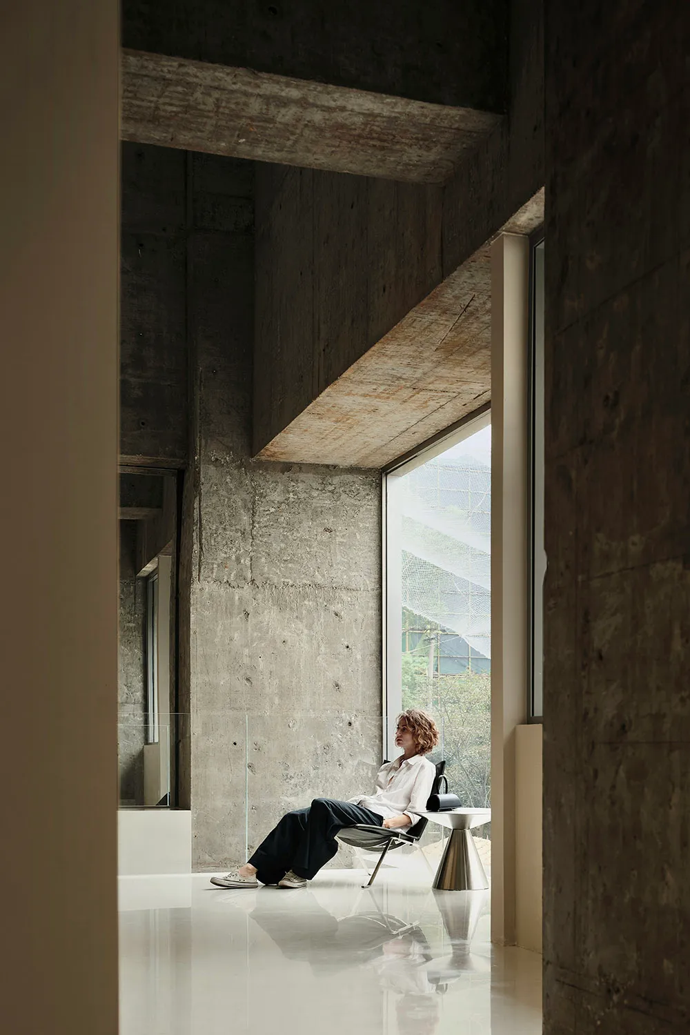 A person sitting in a minimalist interior space with concrete walls and a large window.
