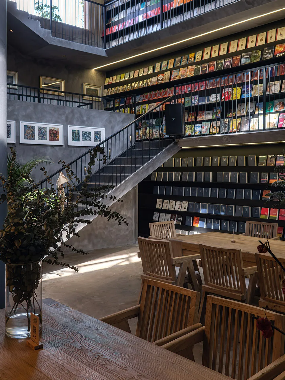 A modern industrial library interior with wooden tables, metal railings, and glass shelves filled with books.