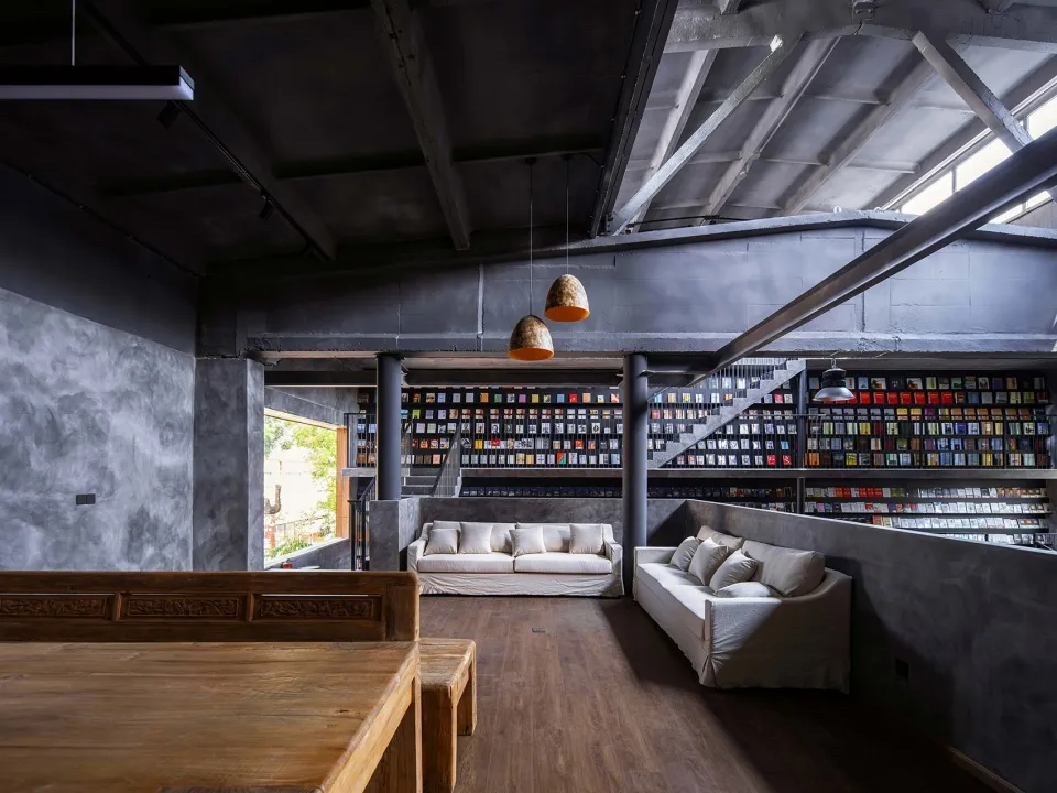 A modern industrial-style library with exposed concrete walls, a staircase, and numerous books on shelves.