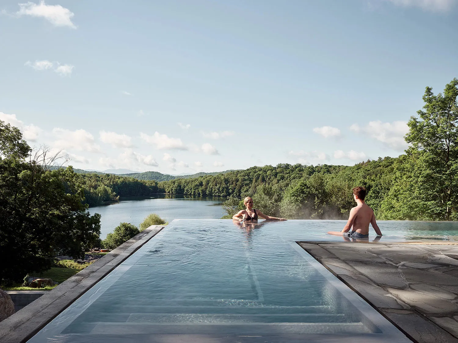 Two people relaxing in a modern outdoor infinity pool overlooking a serene lake and forested hills.