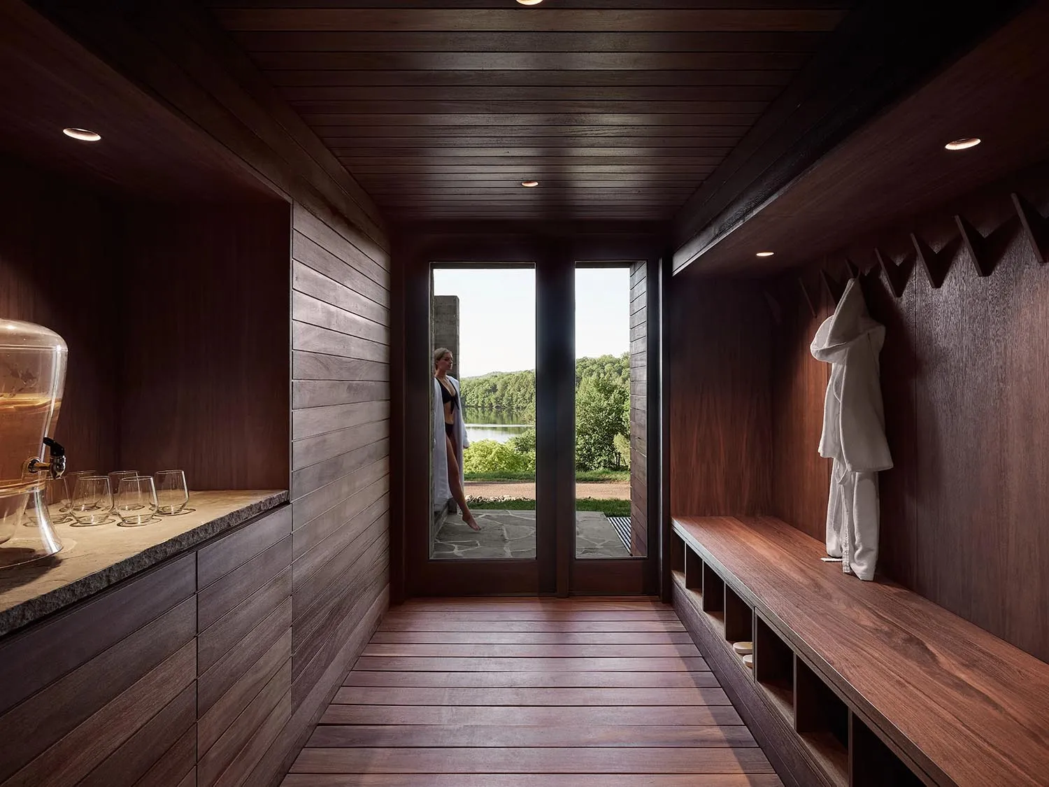 Modern spa interior with wooden walls, floor, and benches, featuring a glass door leading to outdoor greenery and a woman in