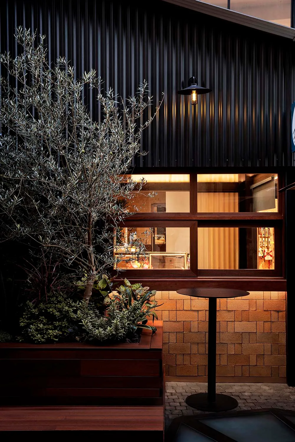 An exterior view of an industrial-style cafe featuring wood planters, brick walls, and metal accents.