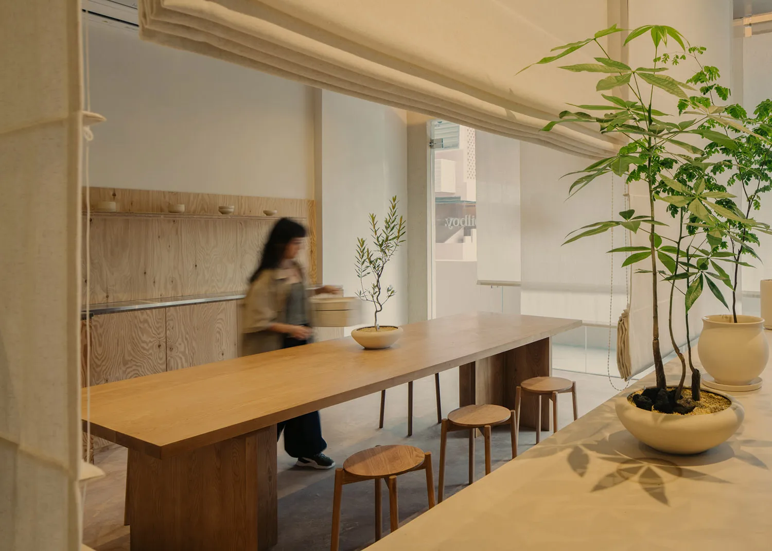 A modern restaurant interior features a long wooden table and stools, with plants and a minimalist design.