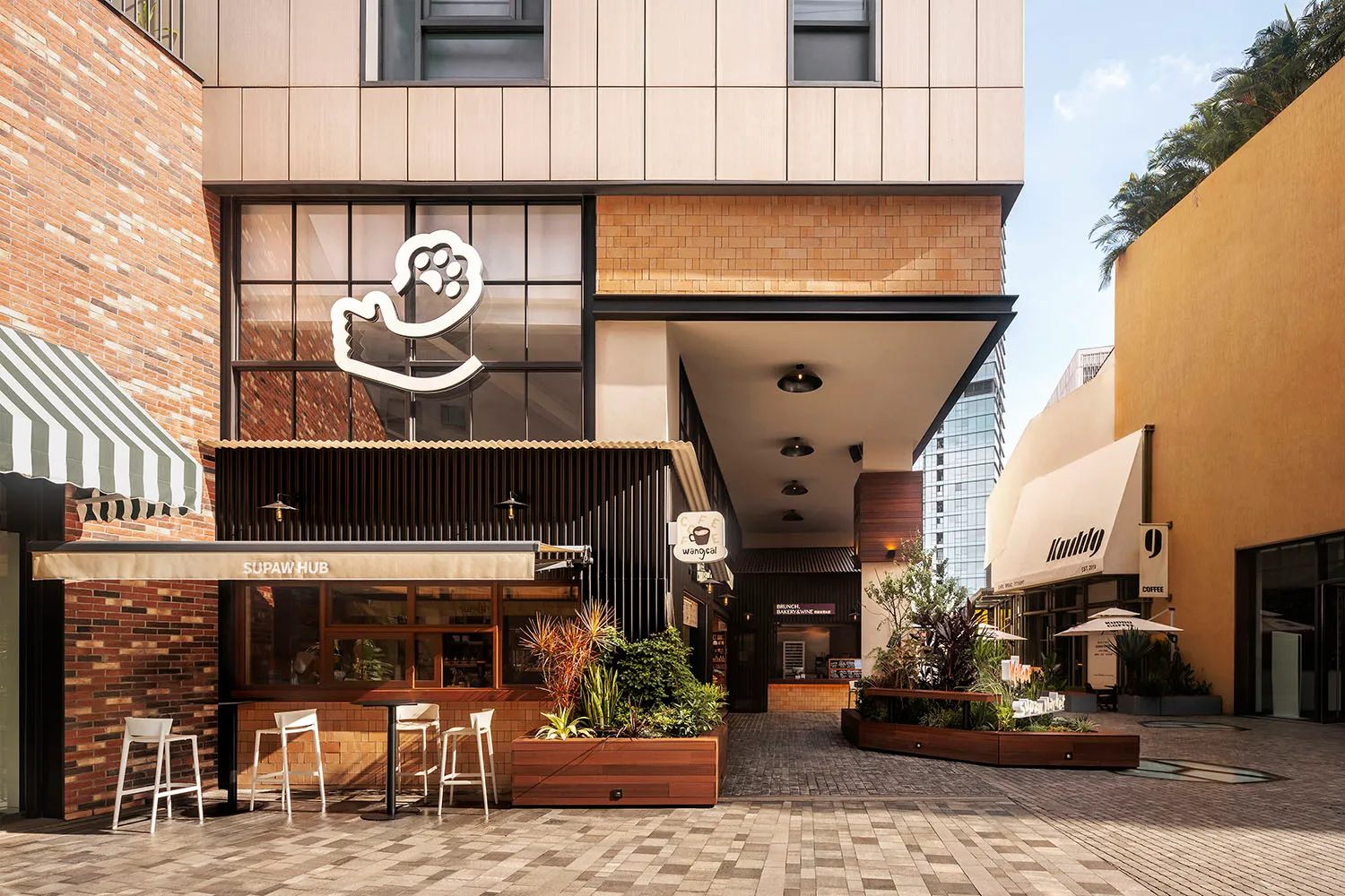 A modern brick storefront with wooden panels and glass windows, featuring potted plants in planters along the walkway.