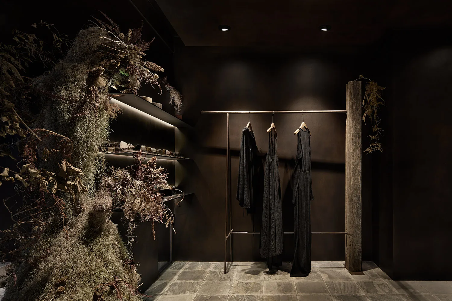 一間採用深色裝潢的現代服裝店,設有木質柱子、擺放植物的層架,以及懸掛衣物的吊桿。