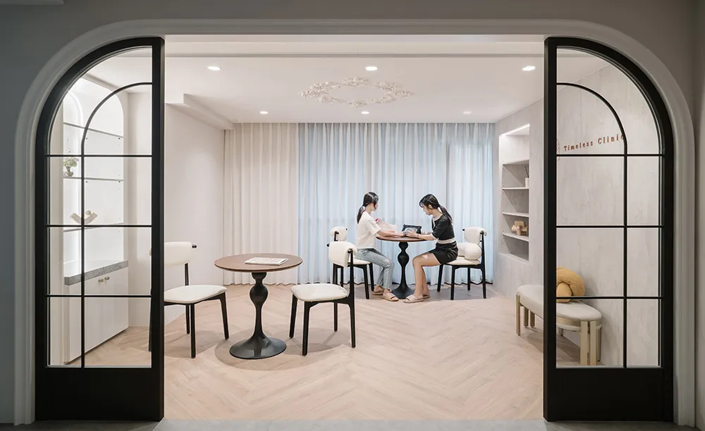 A modern waiting room with light-colored wooden flooring and minimalist decor, featuring two women seated at a table.