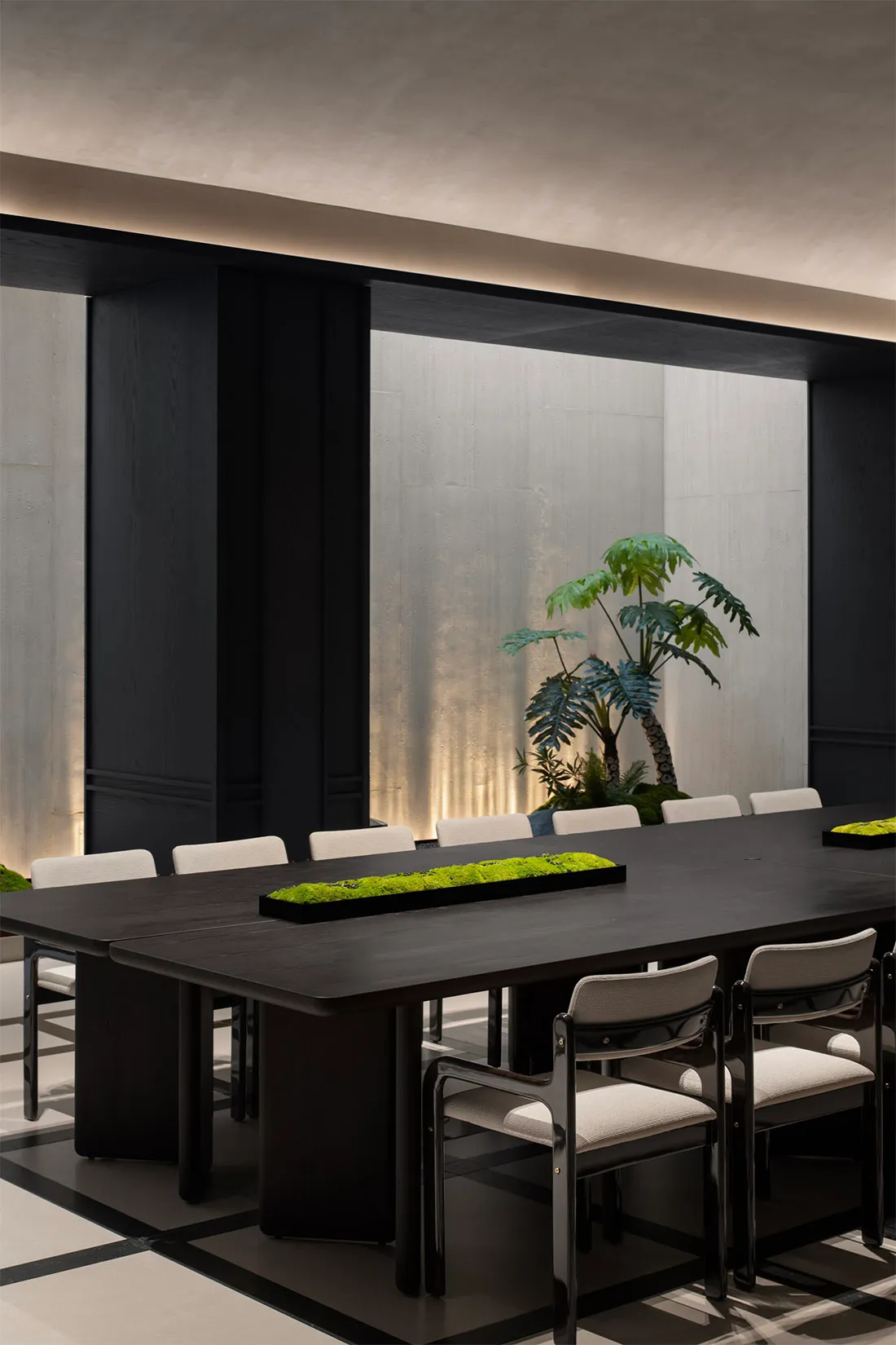 A modern conference room featuring a long black table surrounded by white chairs, with a central green plant arrangement and