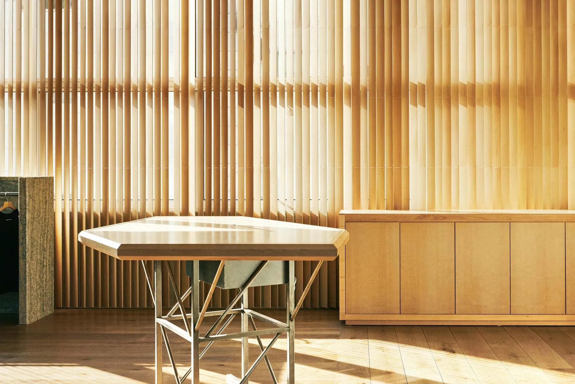 A minimalist office space featuring wooden furniture, with light streaming through vertical blinds.