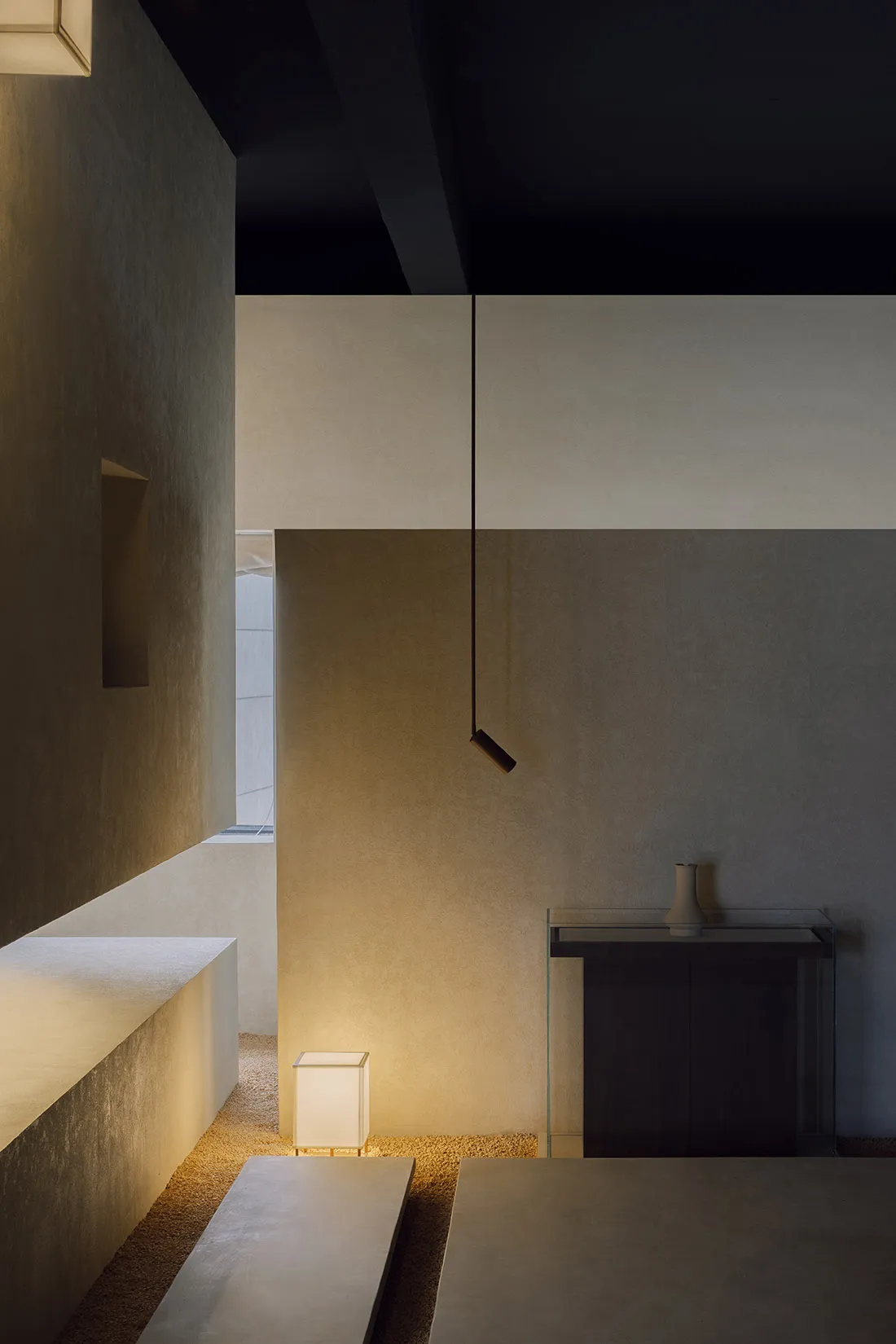 A minimalist bedroom with concrete walls and floor, featuring a modern lamp and a dark nightstand.