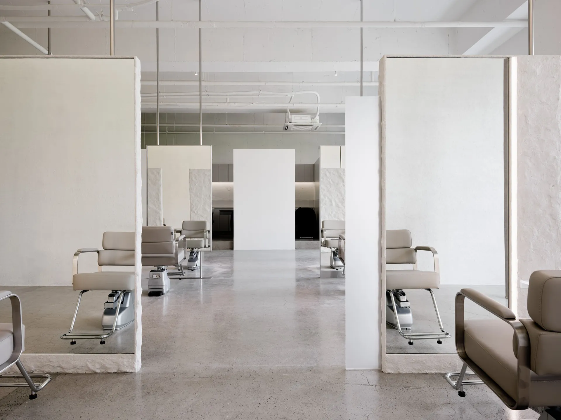 A minimalist hair salon featuring concrete flooring and white walls, with mirrors on the back wall.