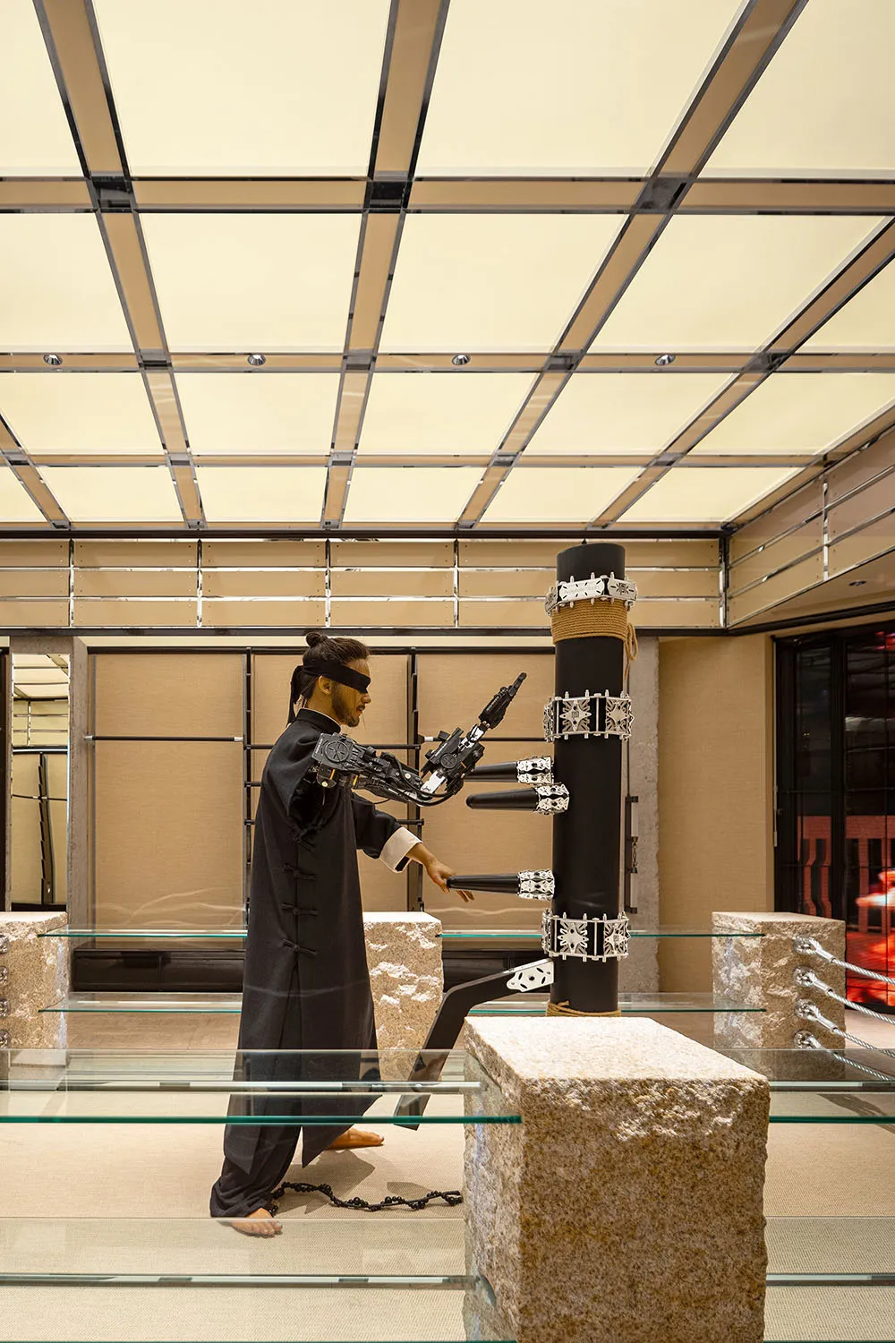 A ninja in traditional clothing and a bladed weapon is performing on a black pole with a blindfold.