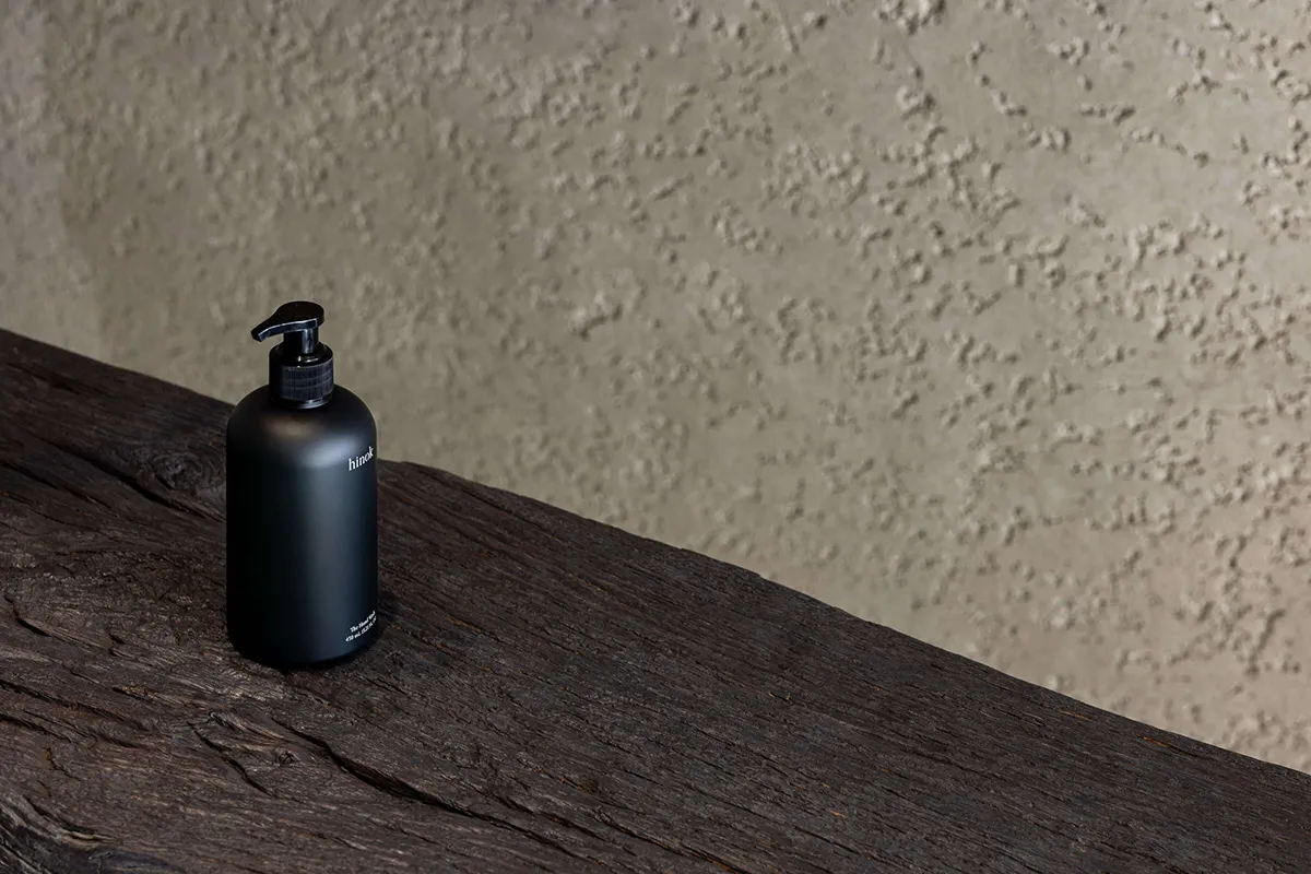 A black metallic bottle with a pump dispenser on a rustic wooden surface and a textured, beige background.