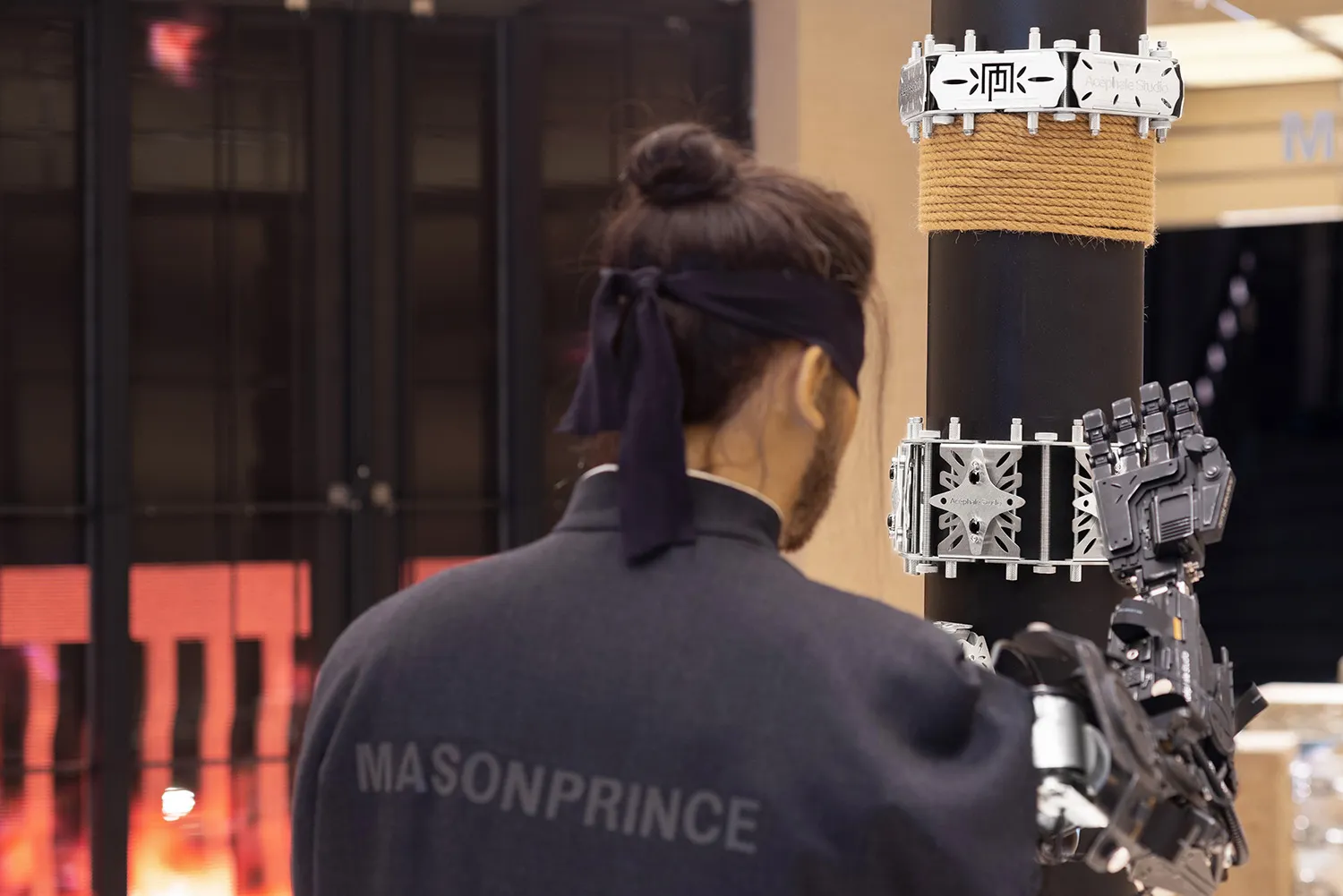 A person wearing a jacket with the words 'MASONPRINCE' on the back, interacting with a robotic arm mounted on a post.
