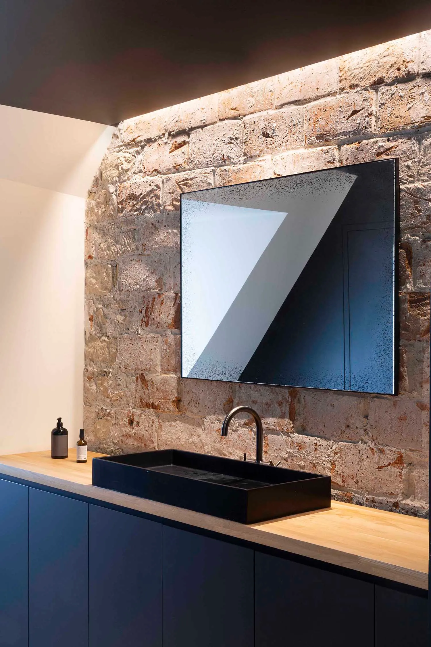 A modern bathroom featuring a black sink and faucet on a wooden counter against a brick wall with a skylight.