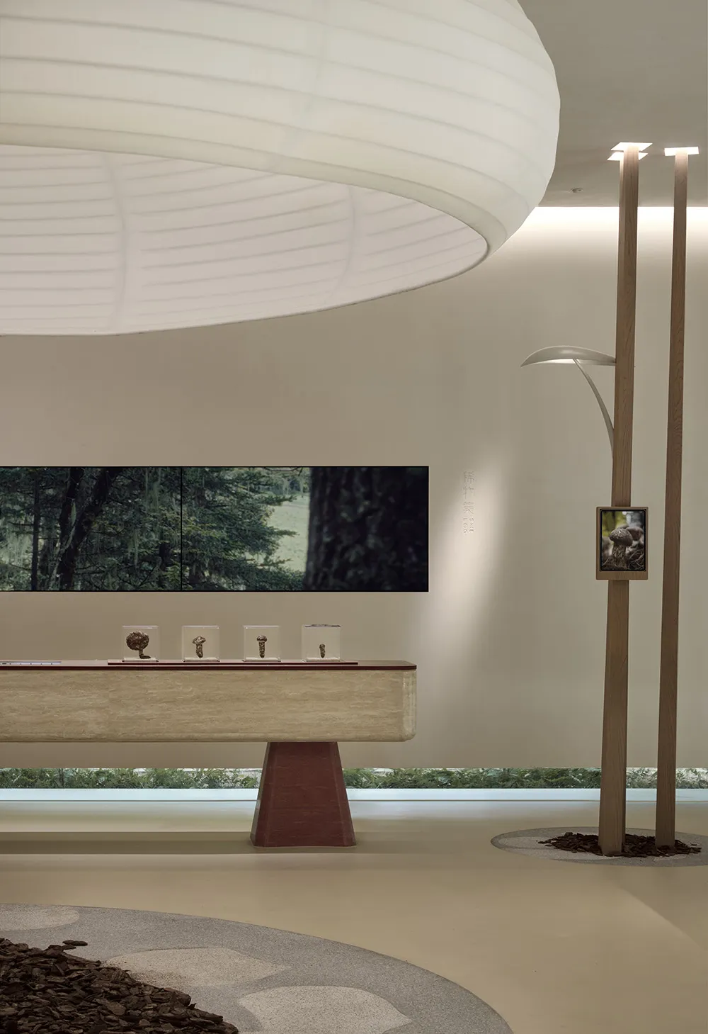 A modern exhibition space featuring a circular light fixture, a wooden table with glass cases, and digital screens displaying