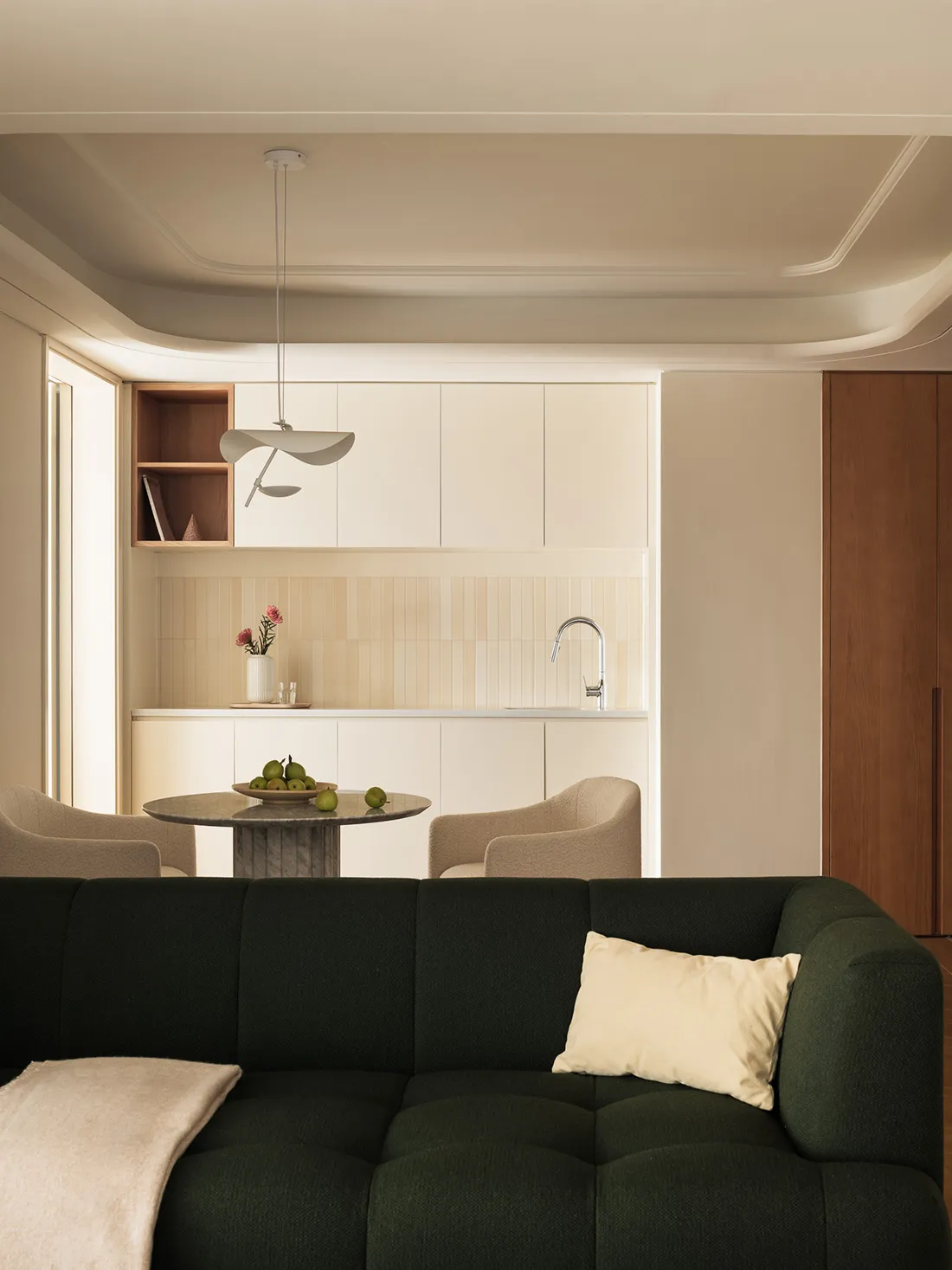 A modern living room featuring a green sofa, beige chairs, and a kitchenette with a sink and open shelving.